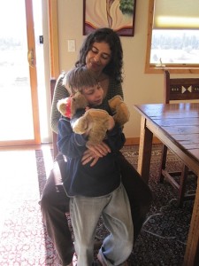 A mother holding her son with a stuffed animal next to a kitchen table. 